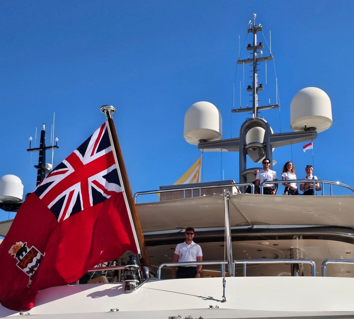 Clos up image of the back of a white yacht. 3 of Oceanskies' team members are stoof on the top deck looing at the camera whie another is stood on the deck just below next to the vessels' registered flag.